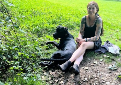 ina allard naturwärts sitzt lachend mit hund leni auf einer grünen wiese